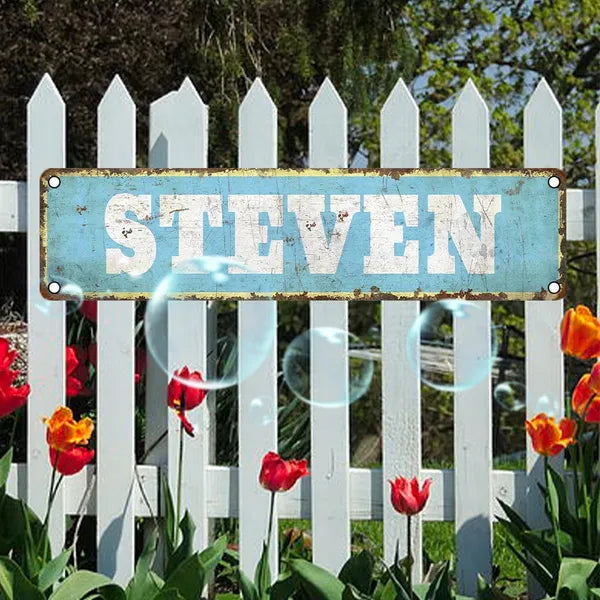 Vintage aluminum name plate sign reading STEVEN on a white picket fence.