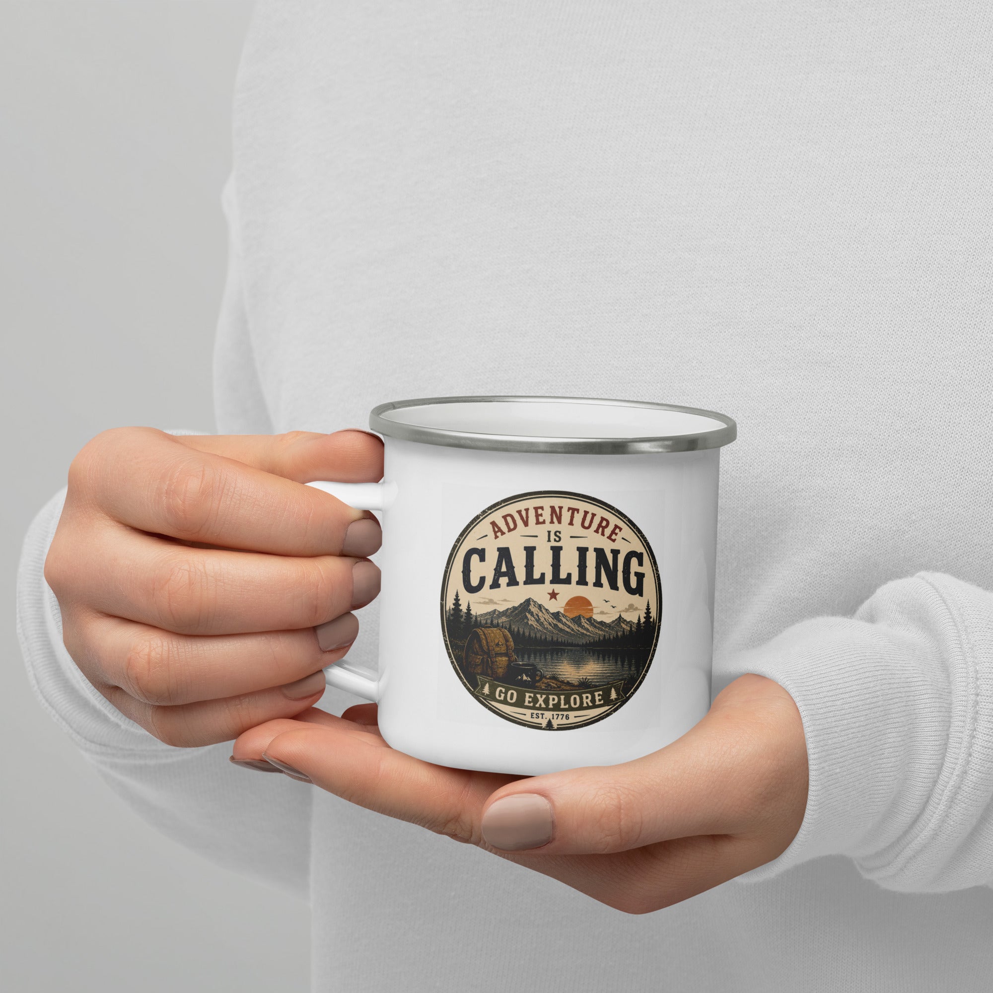 White enamel mug with silver rim held by hands, showing “Adventure is Calling” logo.