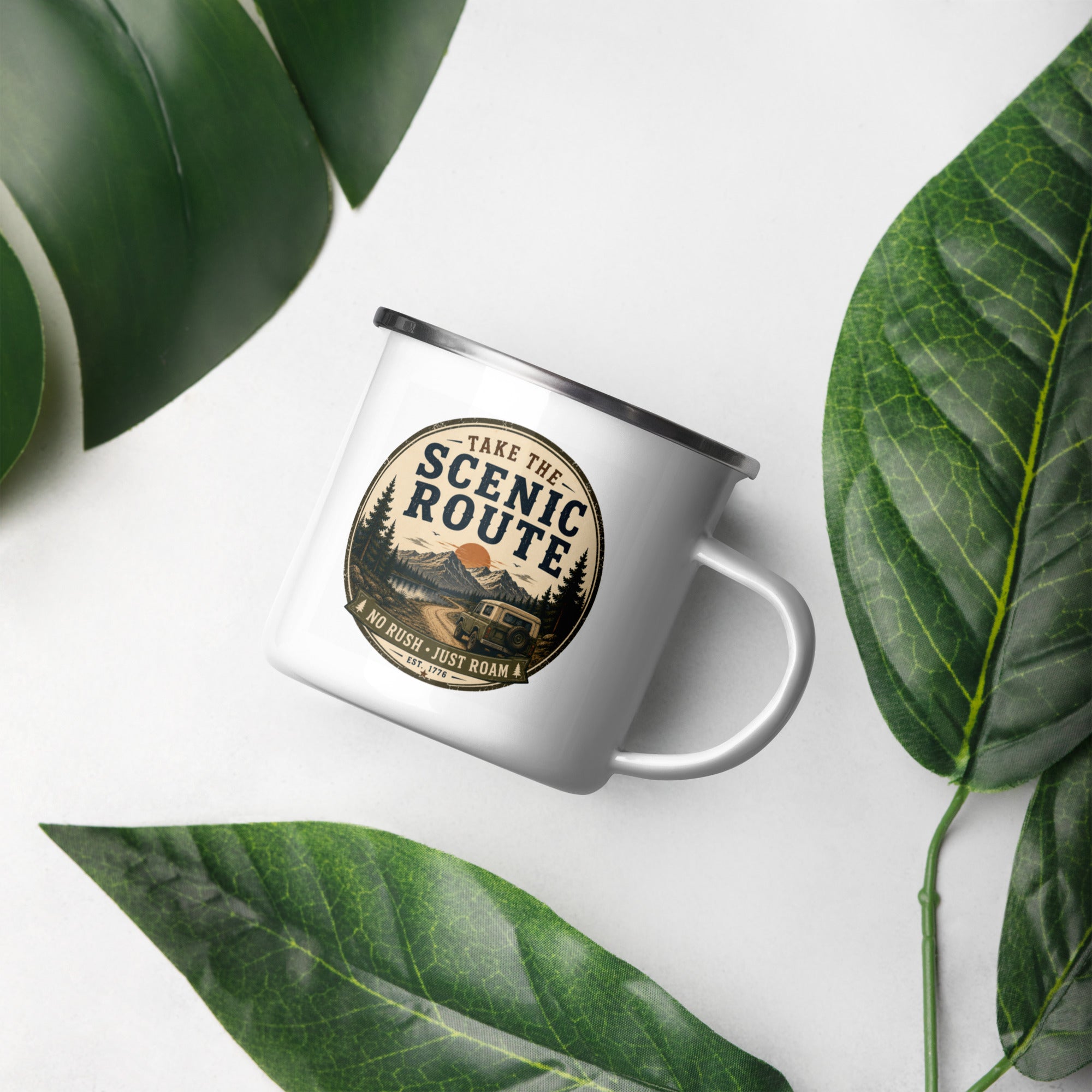 White enamel mug with Scenic Route logo, silver rim and handle, on a leaf background.