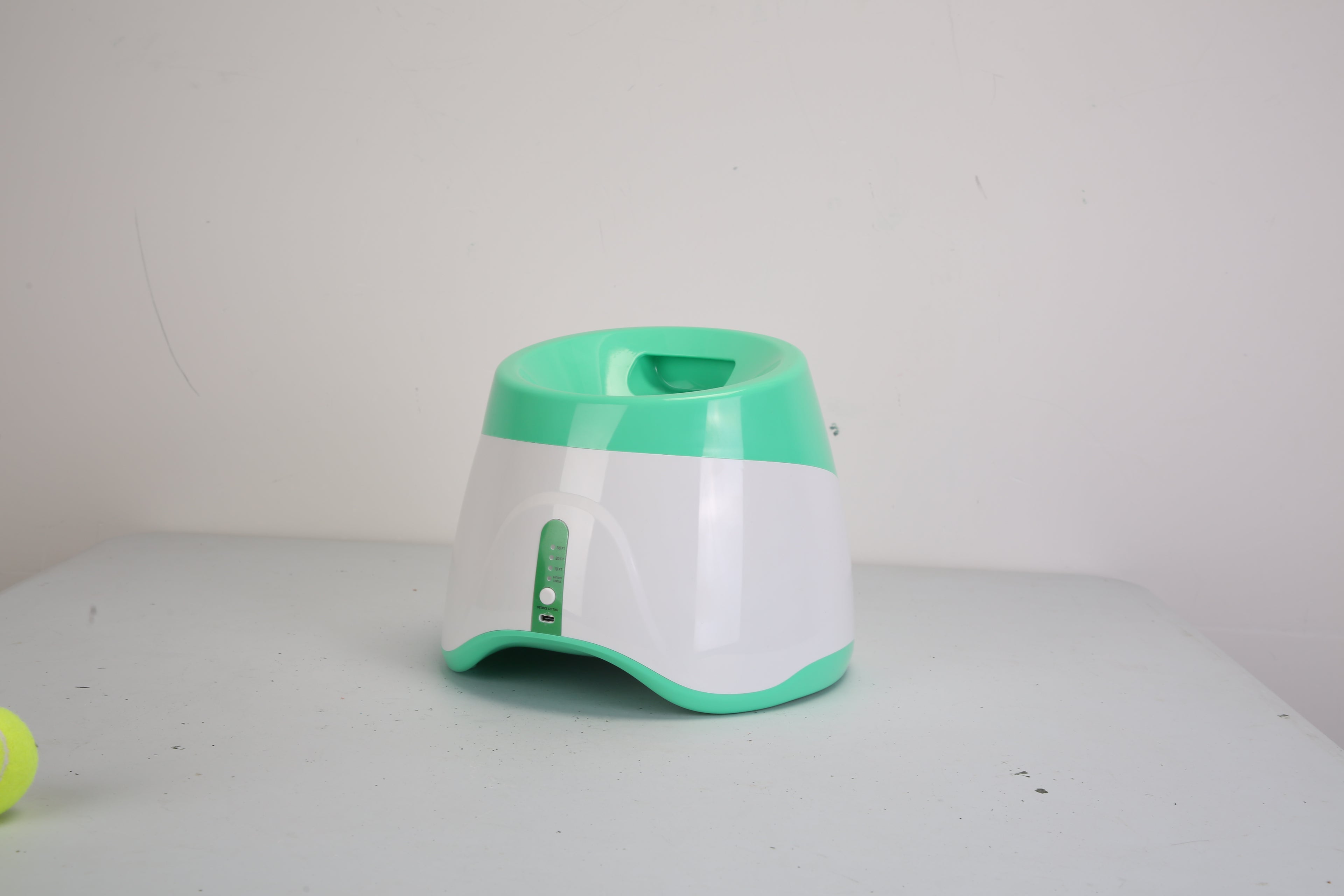 Green and white automatic dog ball launcher with front chute on a tabletop