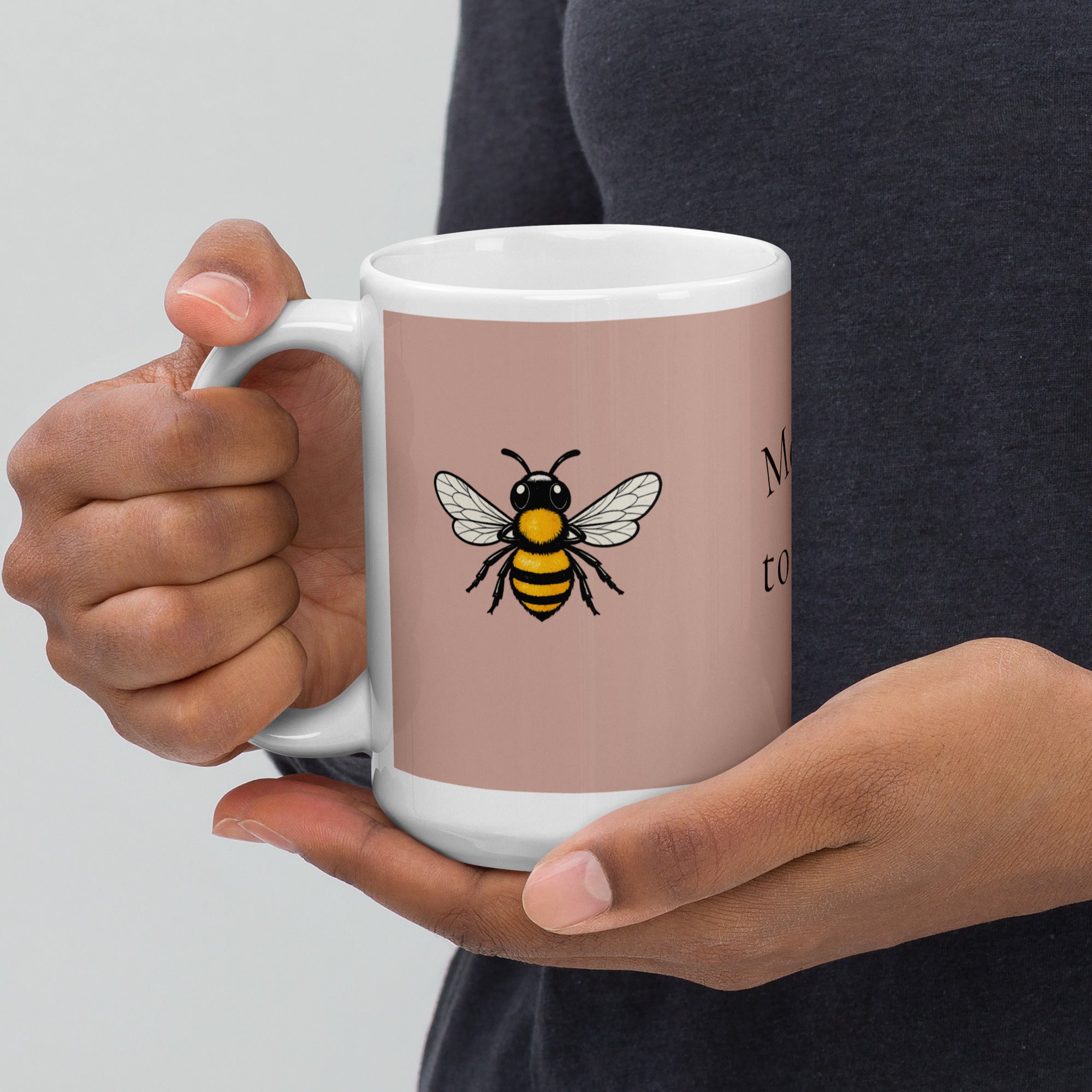Dusty rose ceramic mug with white rim and handle, gripped by hands; bee illustration.