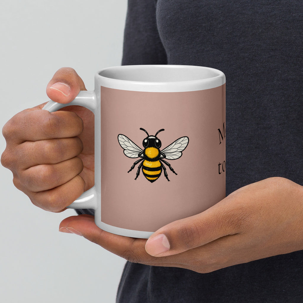 Dusty rose ceramic mug with white rim and handle, close-up of hands holding; bee illustration.