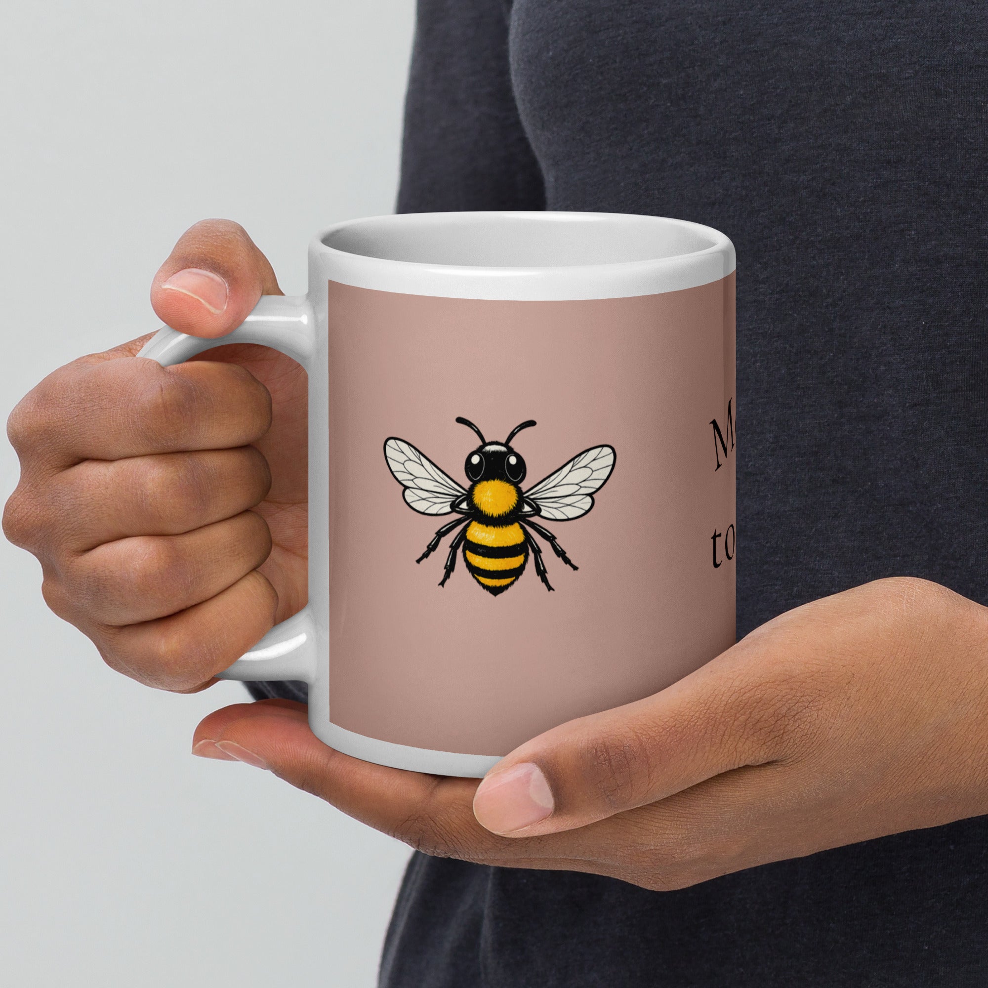 Dusty rose ceramic mug with white rim and handle, close-up of hands holding; bee illustration.